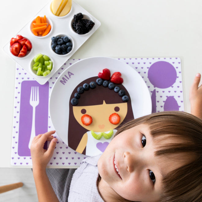 Little girl looking up and using her Dylbug Personalized Little Me Girl Face Dish Set in purple, including  plate, bowl, and placemat - add fruits to her plate to creat funny food faces and make mealtimes fun!