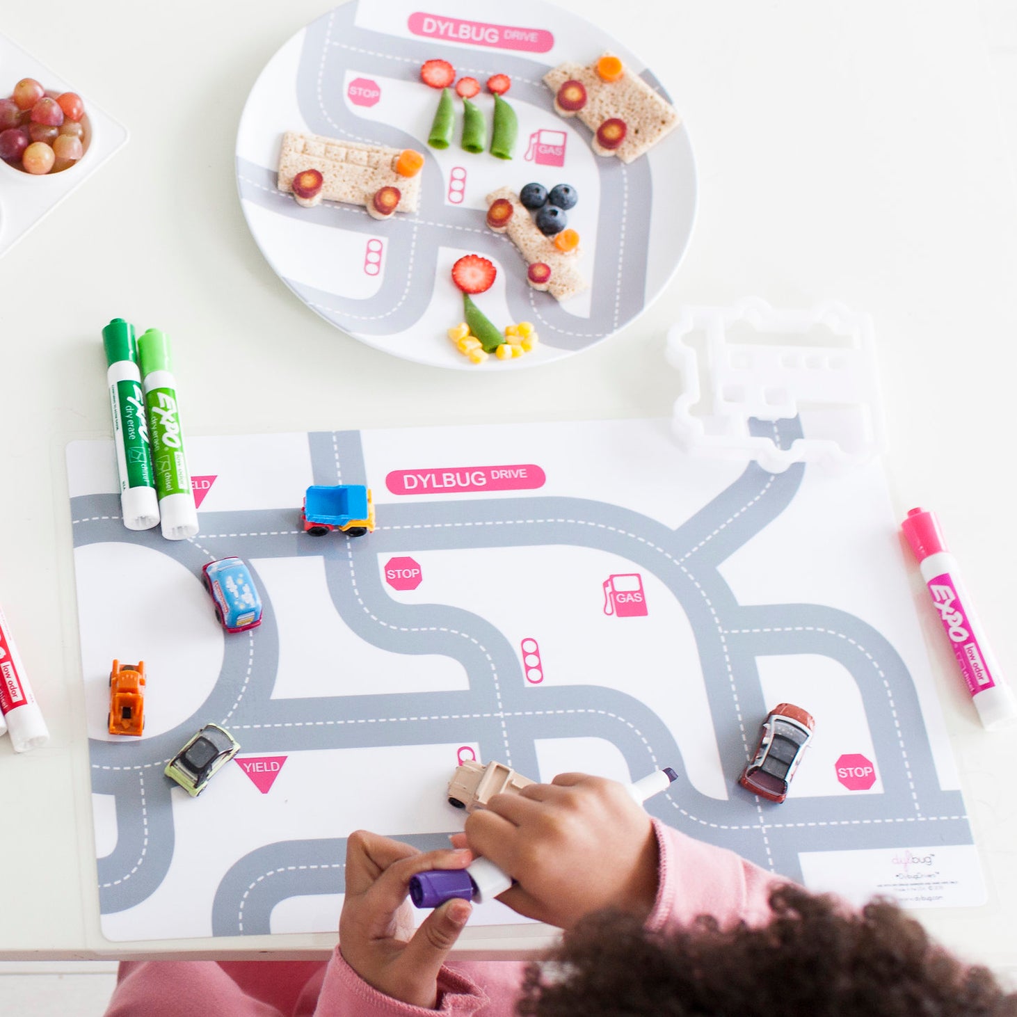 Little girl playing with her custom Dylbug Cars placemat in pink from our Drivers Collection. Next to her car plate with sandwich cars made from our custom vehicle food cutter.