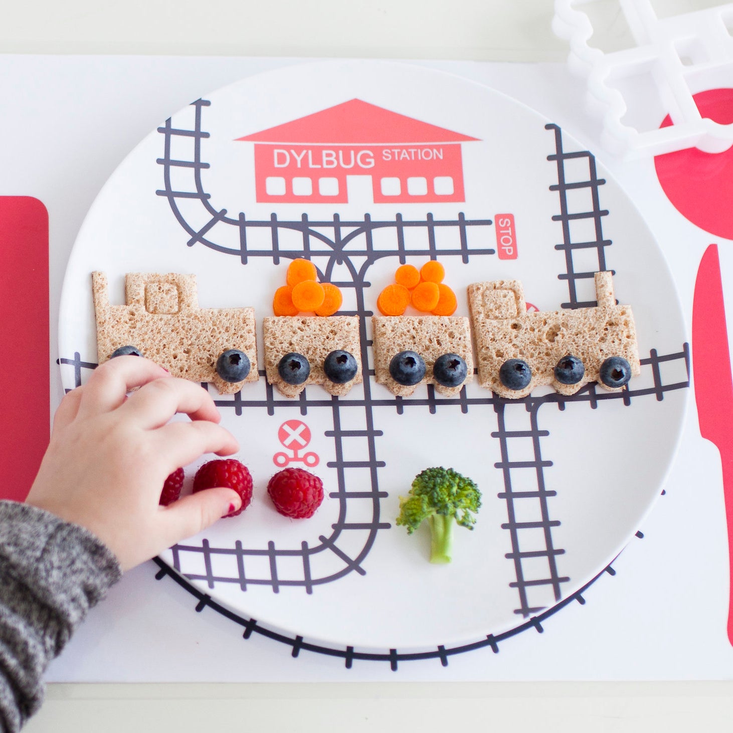 Kid's hand playing with their Dylbug Train plate in pink from our Drivers Collection. Using train food cutter to make a full train sandwich. Perfect for the active train loving kid