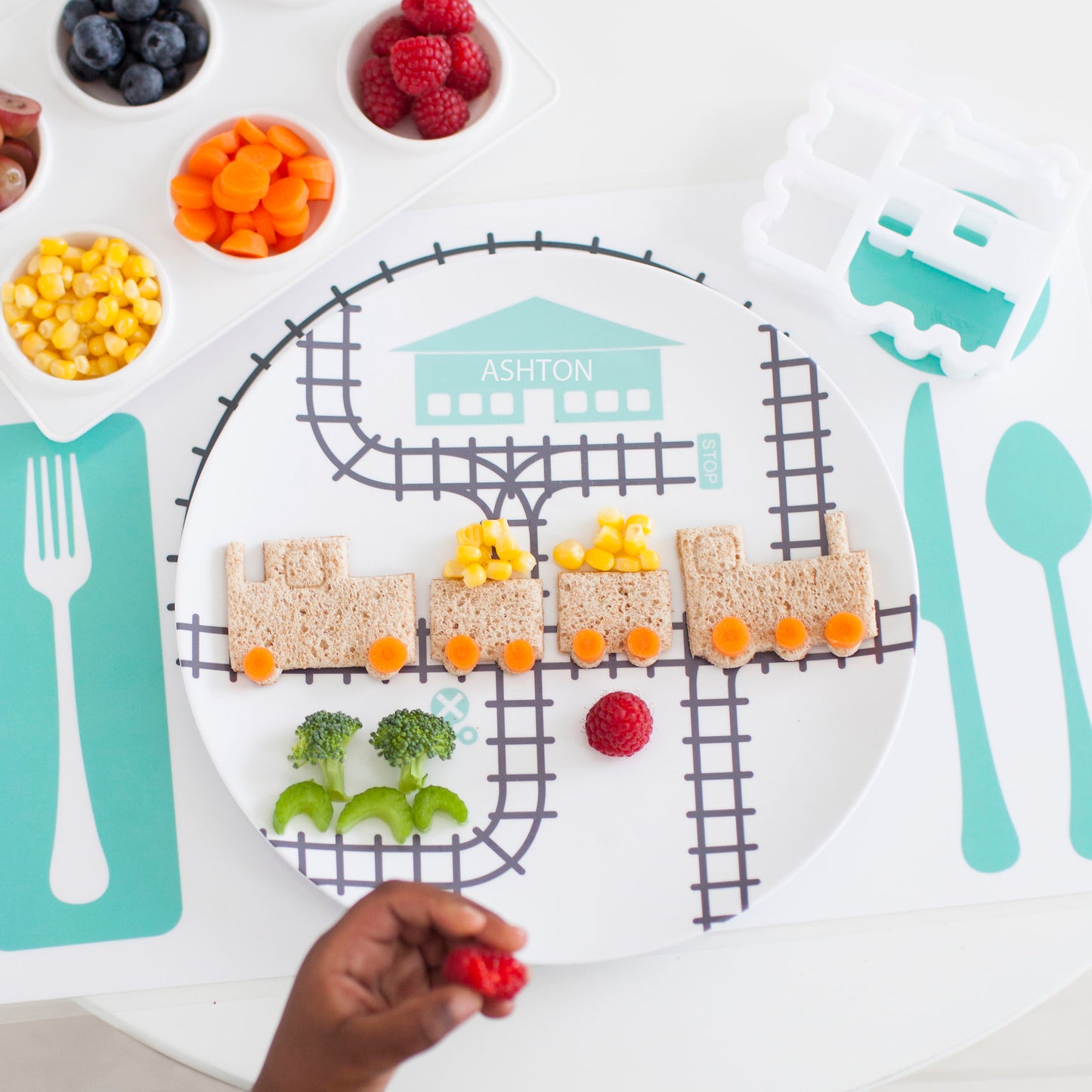 Kid's hand playing with their Dylbug Train plate in teal from our Drivers Collection. Using train food cutter to make a full train sandwich. Perfect for the active train loving kid