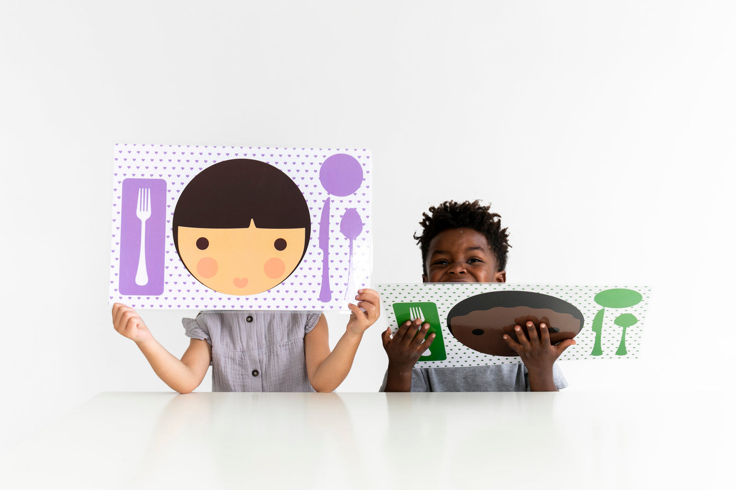 Little girl holding up her Dylbug personalized little me girl placemat in purple. Sitting next to little boy holding his personalized placemat in green
